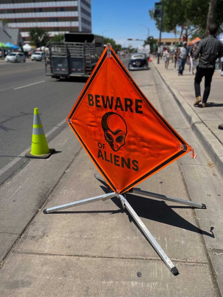 An orange "Beware of Aliens" road sign with a photo of an alien sitting off the side of a road
