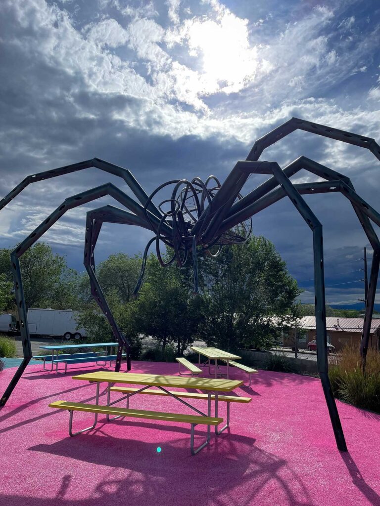 A sculpture of a spider over a couple of tables on pink cement