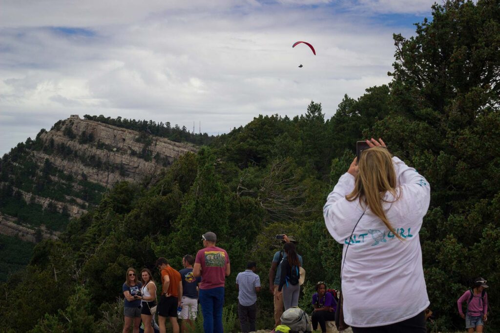 People watch a paraglider fly above a moutain peak