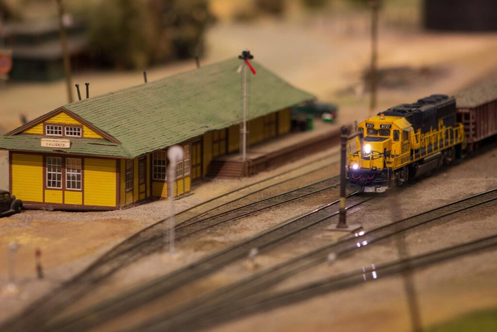 A blue and yellow Santa Fe train running passed a yellow depot on a model train layout