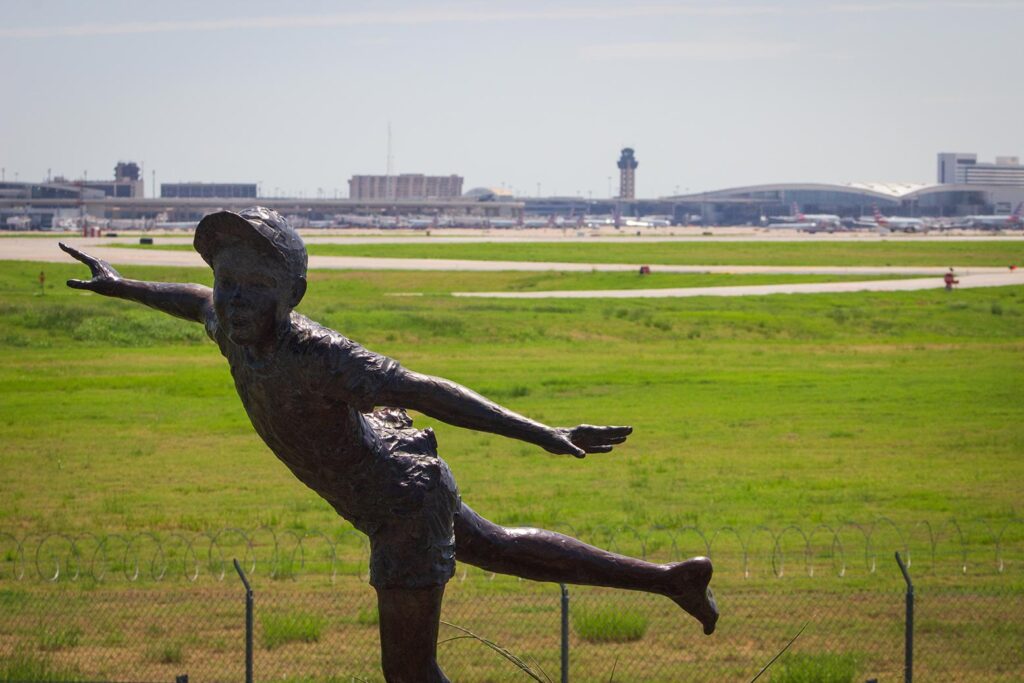 A statue of a boy with his arms spread like an airplane with an airport in the background