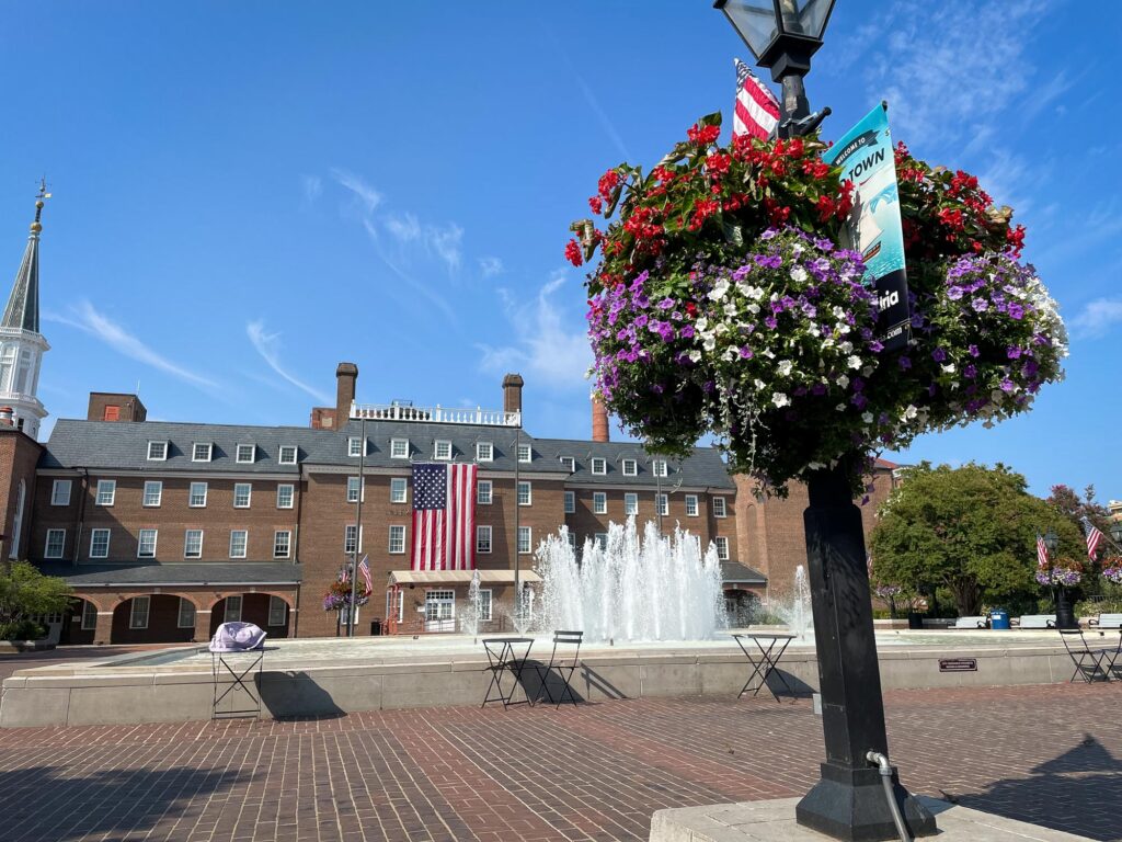 A building with a large, vertical American flag with a flagpole wrapped in flowers in the foreground and a water fountain behind it