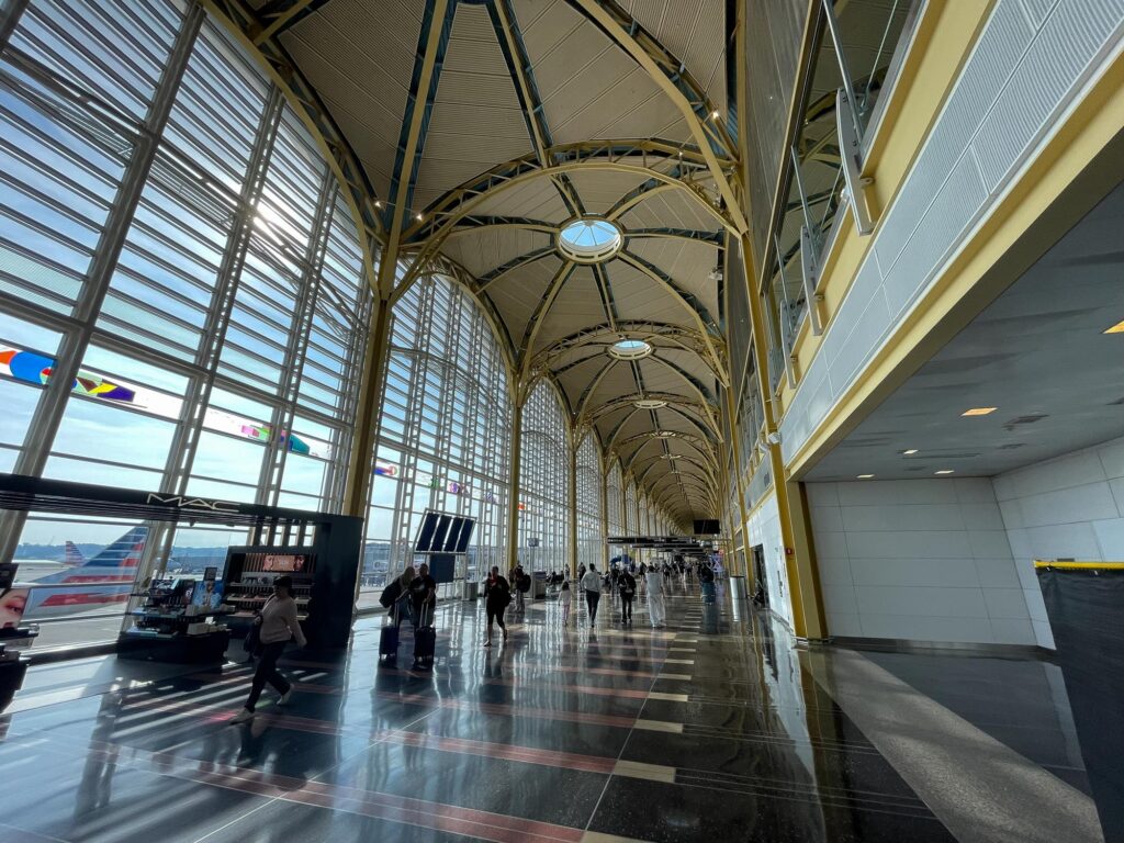 A large walkway at an airport with a high, gold ceiling and large windows on the right