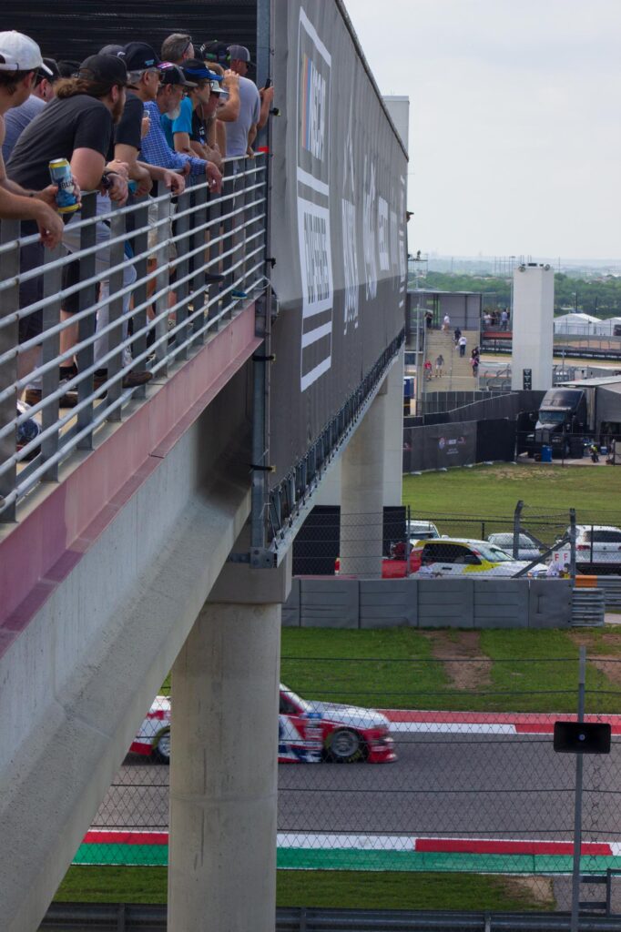 People watching a race from a pedestrian bridge with a race truck driving beneath it