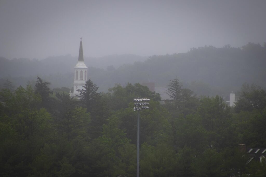 The steeple of a chapel over top green trees and viewed from a distance