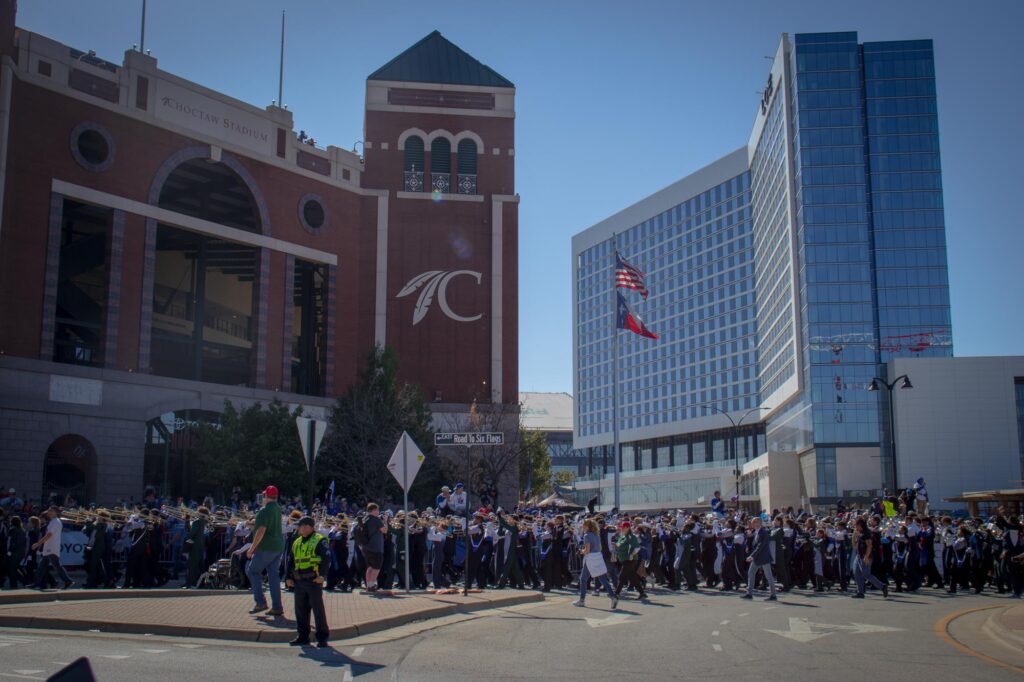 A marching band in navy blue and white uniforms marching in a parade outside a baseball stadium