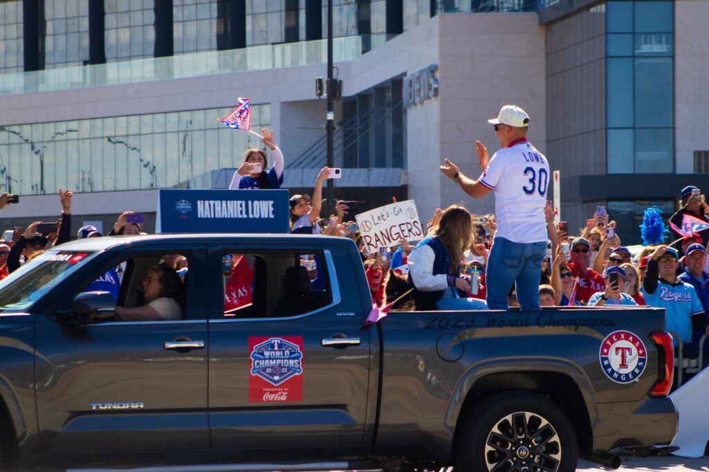 A man in a Texas Rangers jersey standing in the back of a pickup truck in a parade