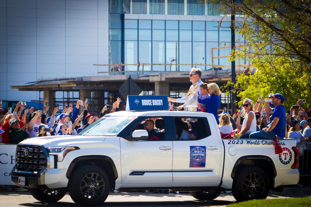 People in Texas Rangers jerseys and jackets in the back of a pickup truck with the World Series Trophy on the roof in a parade