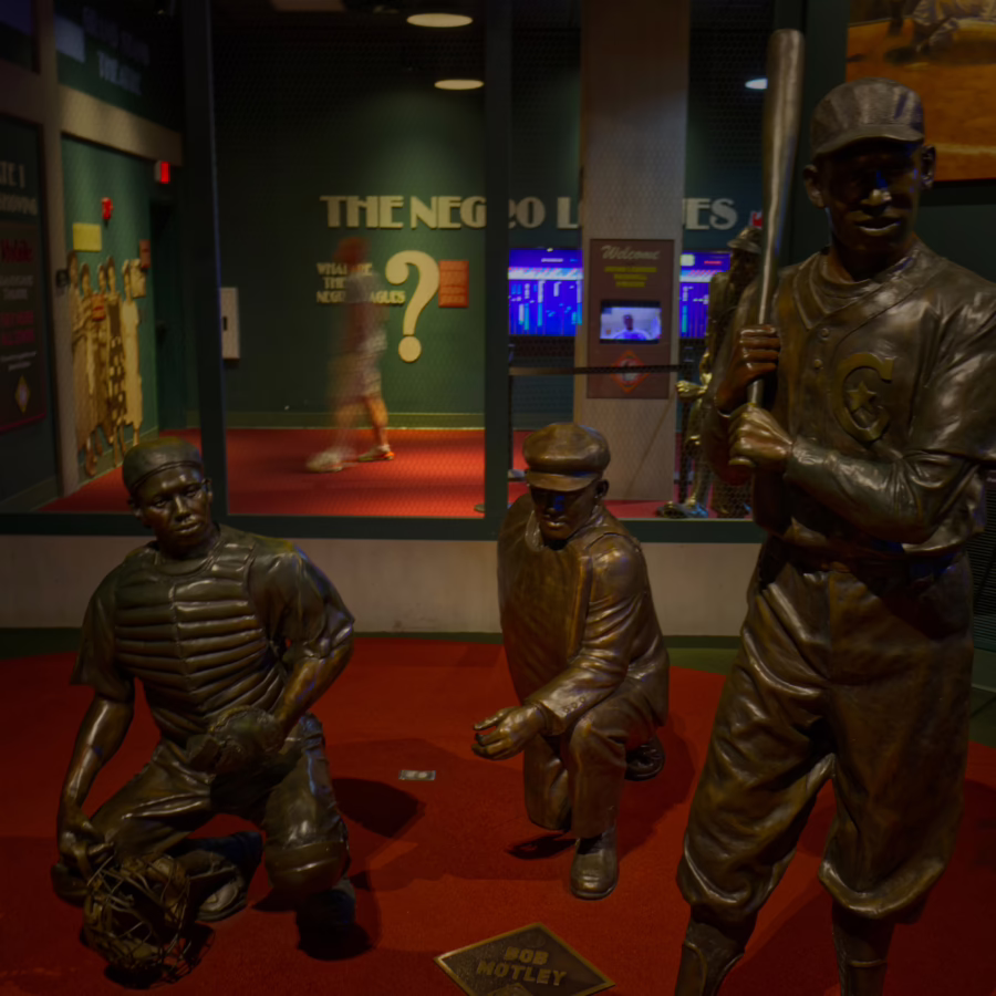 Three bronze statues—a standing batter holding a bat, a kneeling catcher with his mitt, and an umpire in a crouch—are arranged on a red carpet inside the Negro Leagues Baseball Museum.