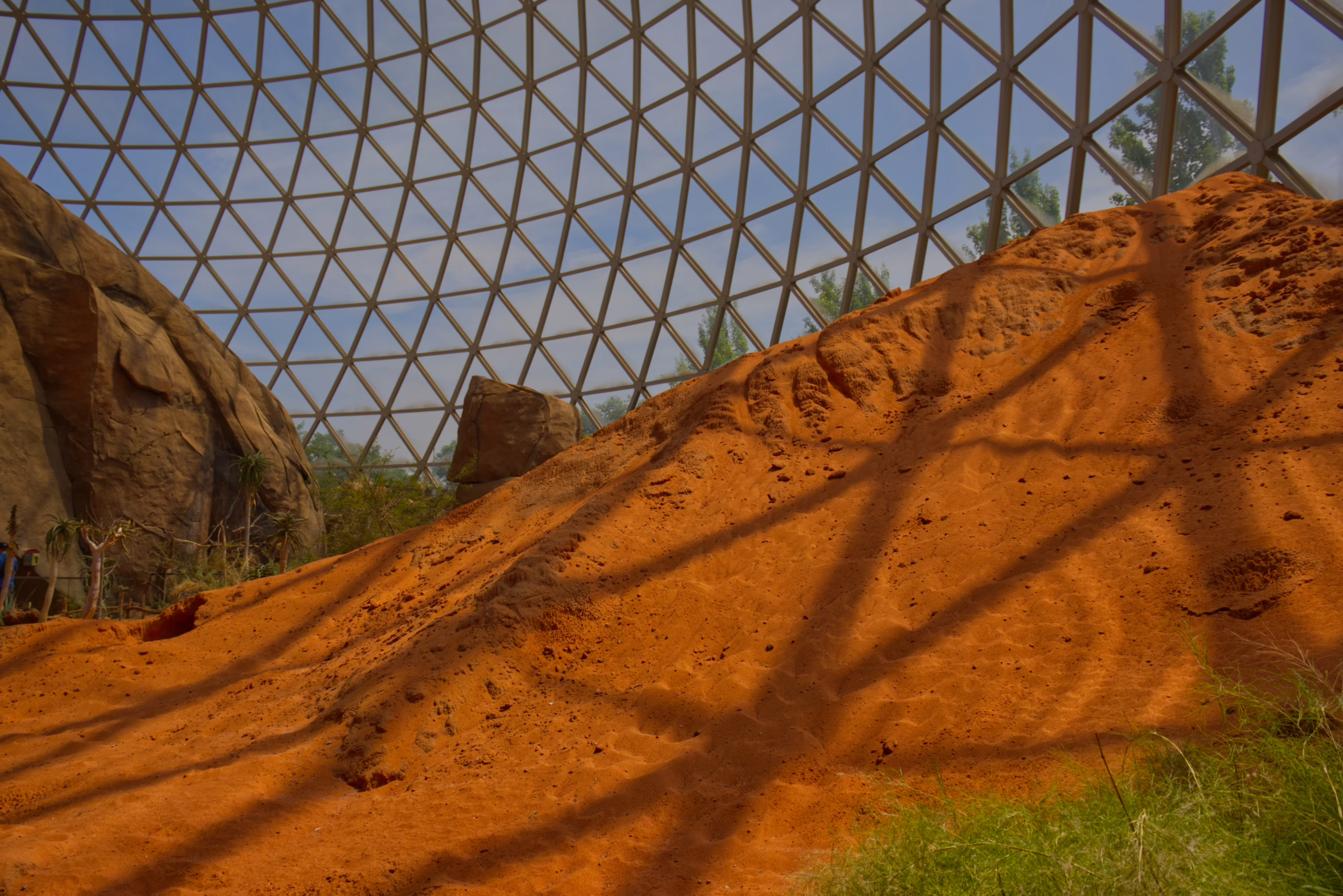The interior of a large geodesic dome, with its geometric frame casting shadows across a vast, sloping hill of bright orange sand that mimics a desert landscape.