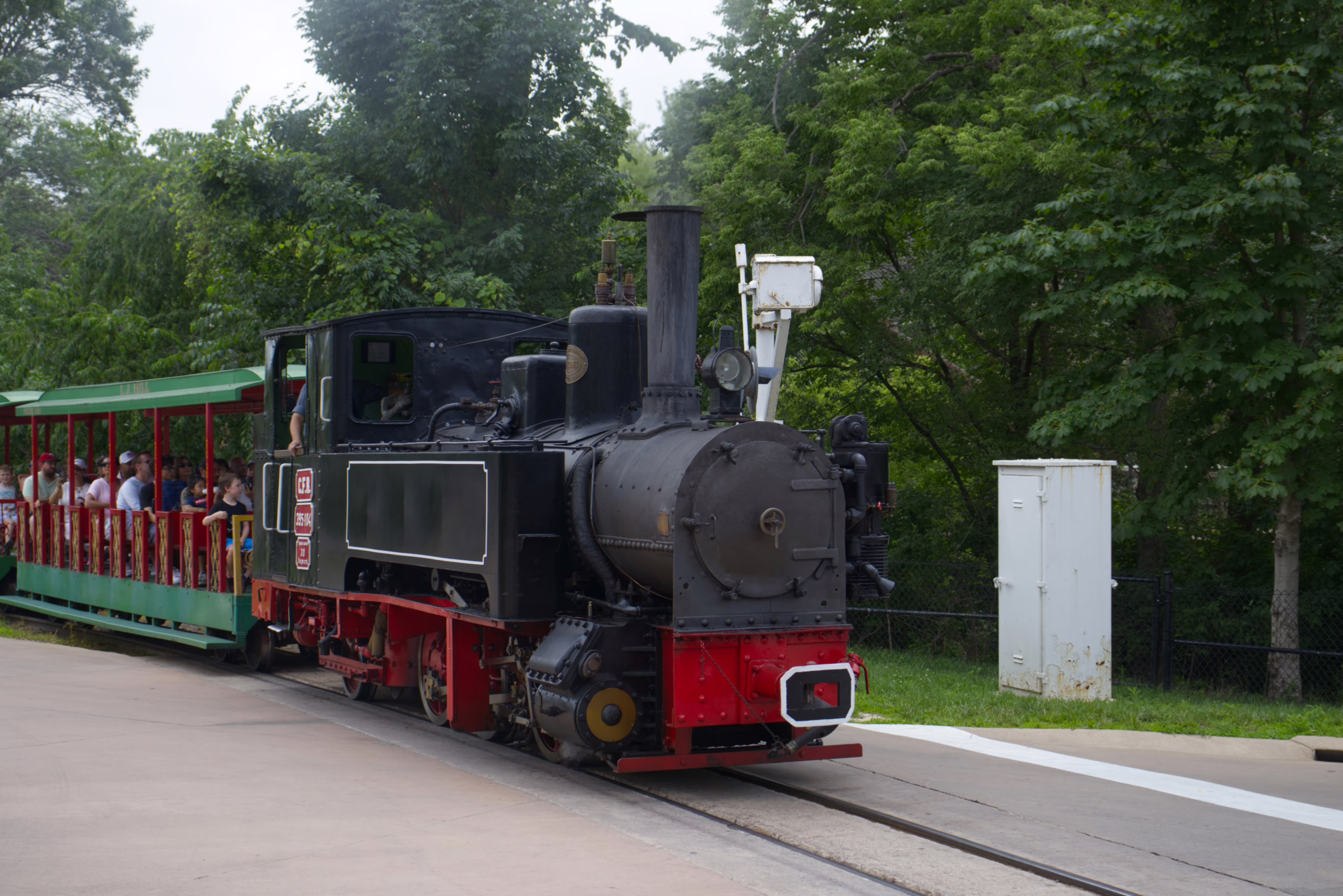 A vintage-style black steam engine with red wheels pulls a green, open-air passenger car full of people along a track at a park or zoo.