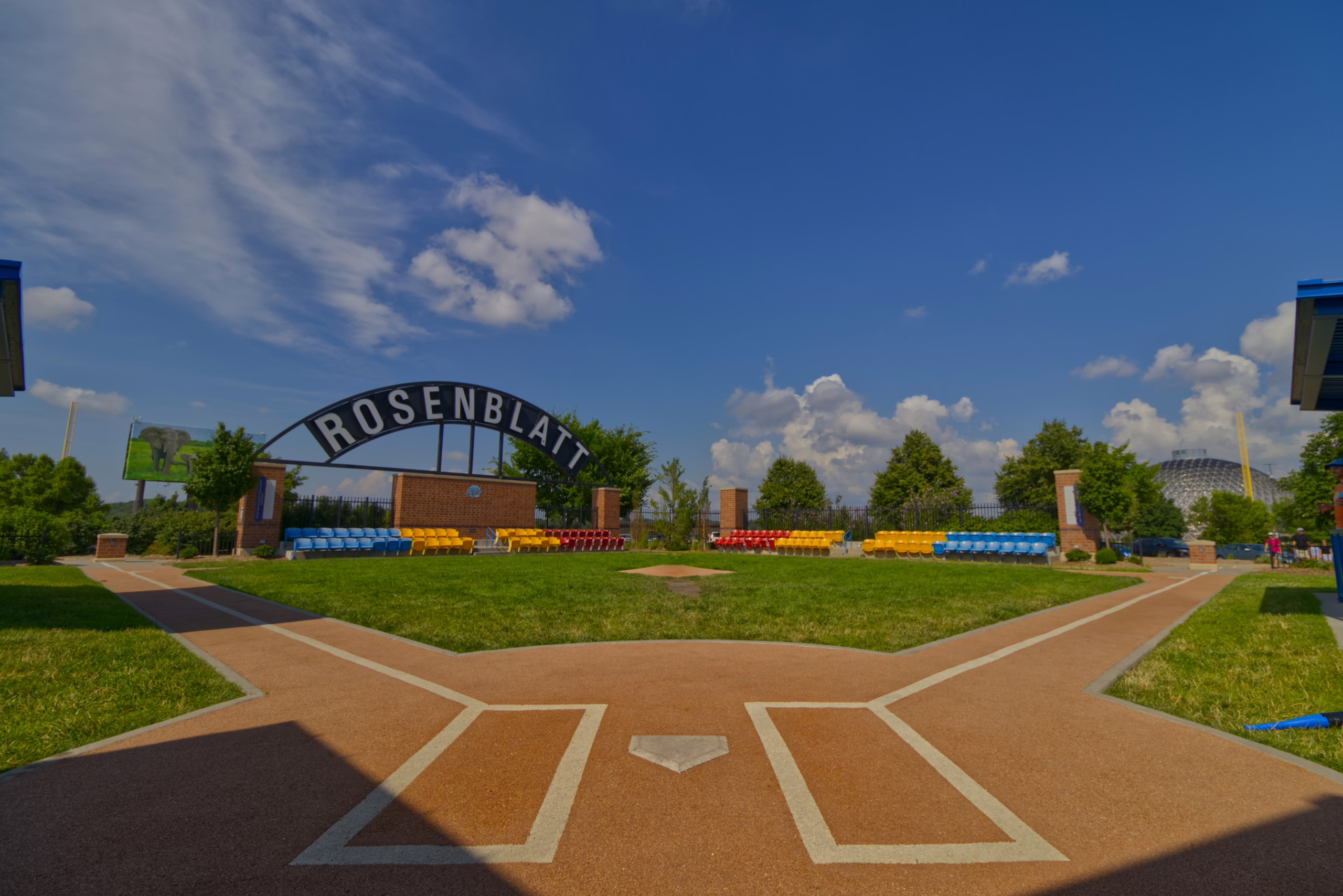 A view from behind home plate of the Rosenblatt Stadium memorial park, featuring colorful stadium seats, a miniature baseball diamond, and the "ROSENBLATT" archway sign under a bright blue sky.