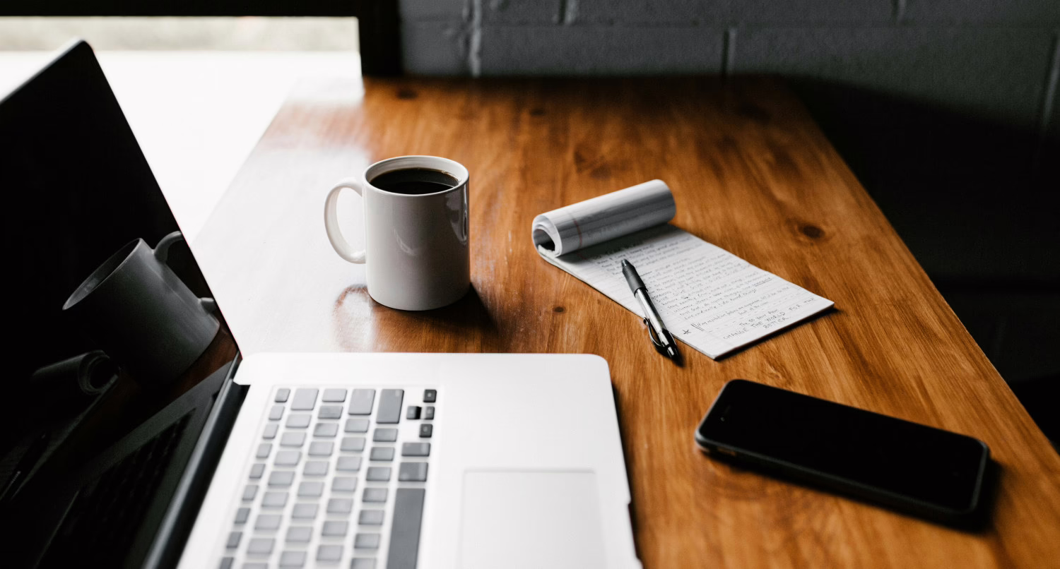 A workspace on a wooden desk featuring an open laptop, a white mug of black coffee, a flipped-over notepad with a pen, and a smartphone.