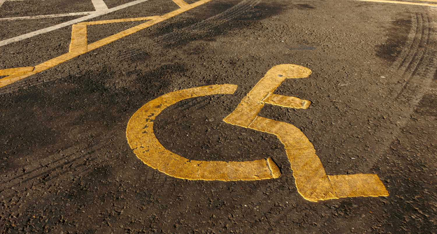 A close-up shot shows a yellow handicapped parking symbol painted on dark asphalt. The symbol, which depicts a stylized figure in a wheelchair, is slightly worn and textured, suggesting age and use. Faint tire marks are visible on the surrounding asphalt. In the background, out of focus, are parts of other yellow parking lines. The lighting highlights the texture of the paint and the road surface.