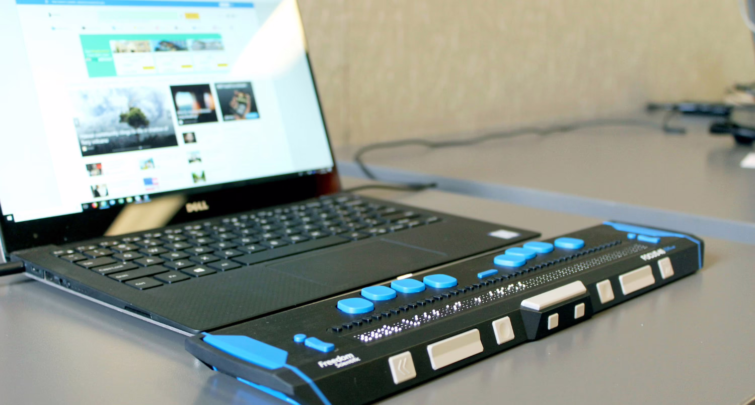 A low-angle indoor shot shows a matte black Dell laptop with a dark keyboard and a bright screen displaying a news website. To the right of the laptop is a black and blue Freedom Scientific braille keyboard.