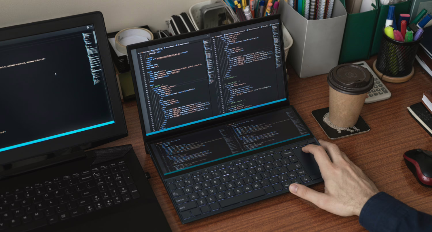 A person's hand rests on the keyboard of an open laptop, which displays lines of code across its screen. Another laptop sits to the left, also open. A coffee cup, pens, and other desk items are scattered around the work surface. A person's hand rests on the keyboard of an open laptop, which displays lines of code across its screen. Another laptop sits to the left, also open. A coffee cup, pens, and other desk items are scattered around the work surface.