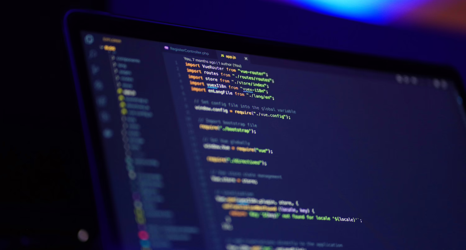 An angled, close-up shot of a laptop screen showing an app.js file in a dark-mode code editor. The JavaScript code includes import statements for Vue.js libraries such as VueRouter and VueI18n.
