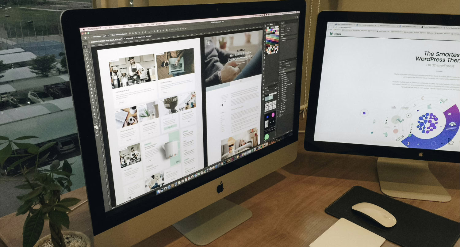 A dual-monitor computer setup on a wooden desk, featuring an Apple iMac on the left displaying Adobe Photoshop with a website design project open, and a second monitor on the right showing a web page about a WordPress theme. A keyboard and mouse are in front of the right monitor, and a small potted plant sits on the left side of the desk near a window with a view of a parking lot outside.