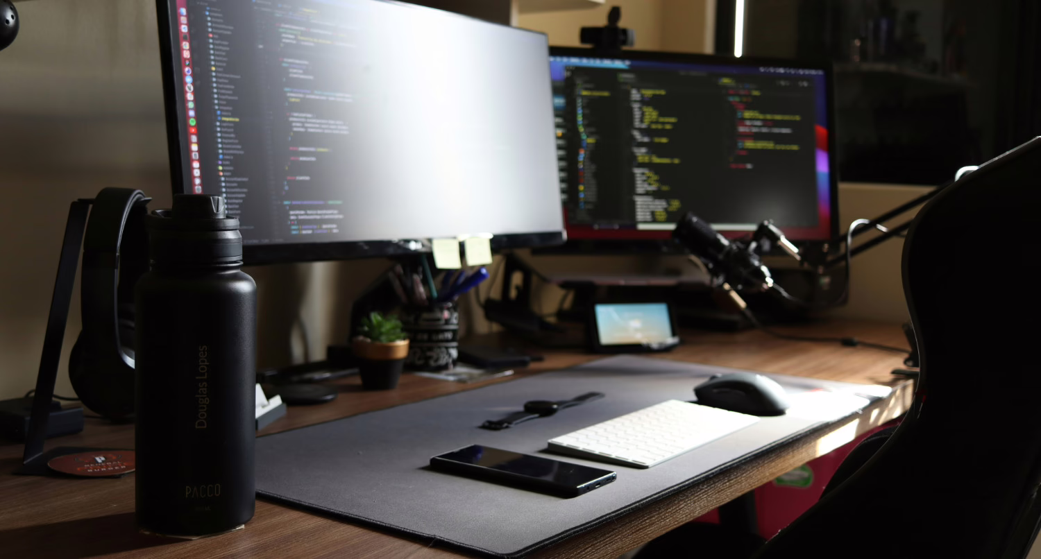 A silver laptop open on a desk displays lines of code within a dark-mode integrated development environment (IDE). A small, orange plush octopus toy sits on the desk to the left of the laptop. In the background, a large screen with blurred colorful shapes is visible.