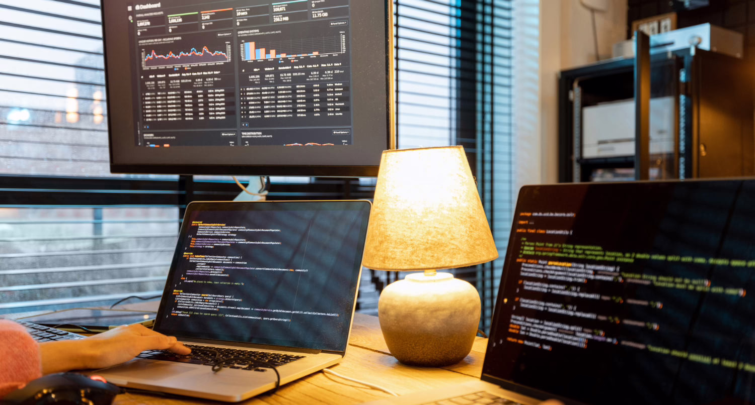 A developer's workspace with three screens displaying code and data. A laptop in the center and a secondary screen on the right show code in dark-themed editors, while a large monitor above displays a dashboard with charts, graphs, and tabular data. A table lamp with a warm glow sits between the screens, and a person's hand rests on the laptop keyboard.