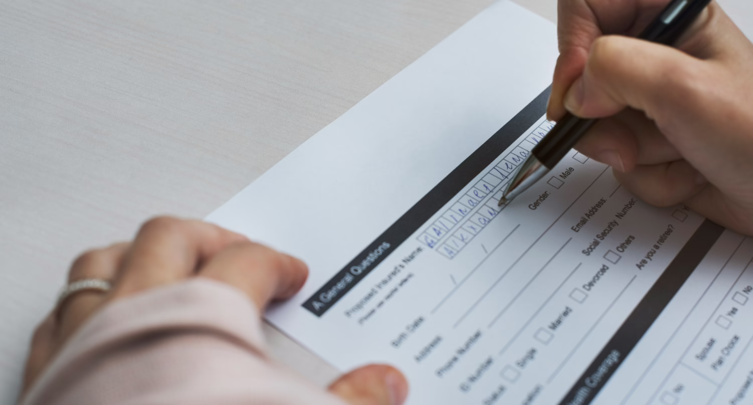 A close-up photo of a person's hands filling out a paper form titled 'General Questions' with a black pen. The form includes fields for name, birth date, address, phone number, email address, gender, and social security number, with some fields already filled in with handwritten text.