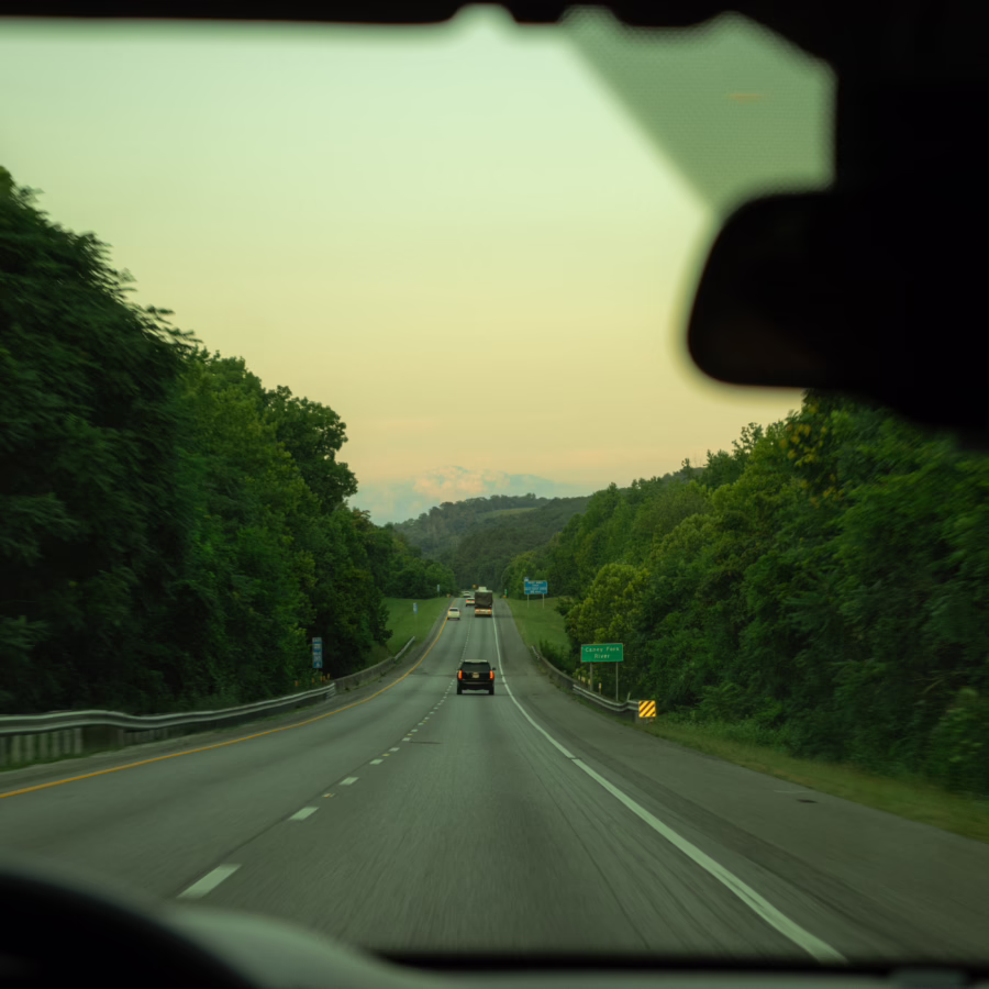View through a car windshield of an interstate winding between forested hillsides at dusk, with a few vehicles ahead, a "Caney Fork River" sign on the right, and a soft yellow-pink sky.