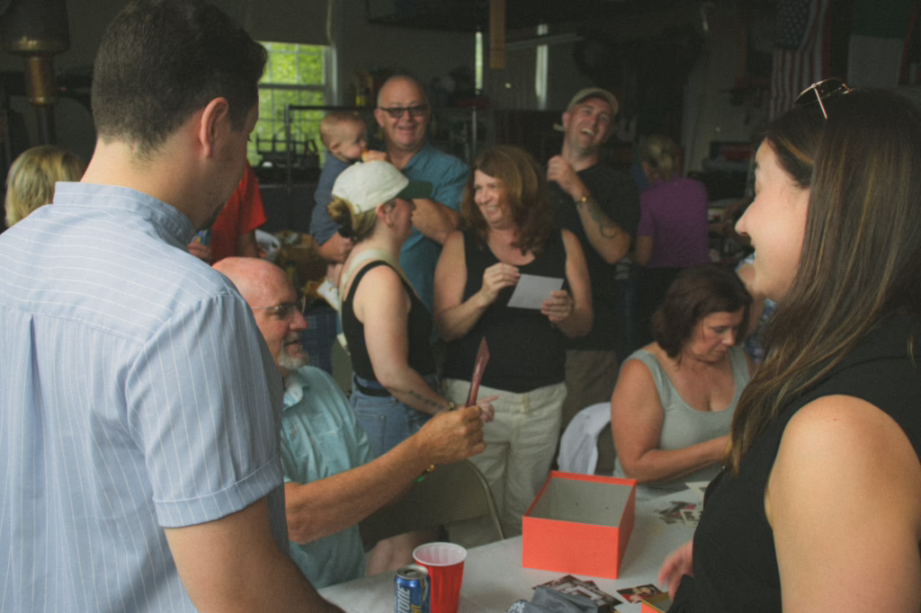 A group of family members gathered around a table inside a garage at the Ciarlo Family Reunion, smiling, talking, and looking through photos pulled from a red box, with drinks and a small American flag visible in the background.